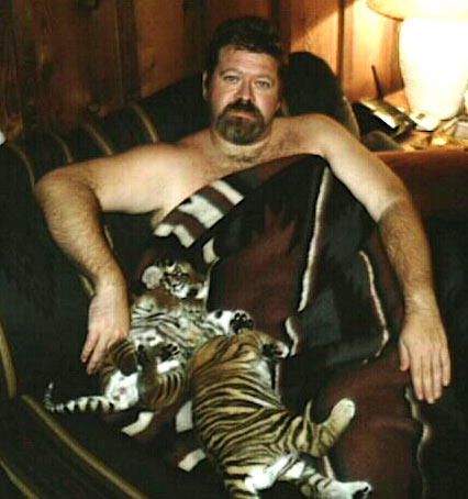 Mike sitting on a sofa with a blanket wrapped around him, snuggling with two small tiger cubs.