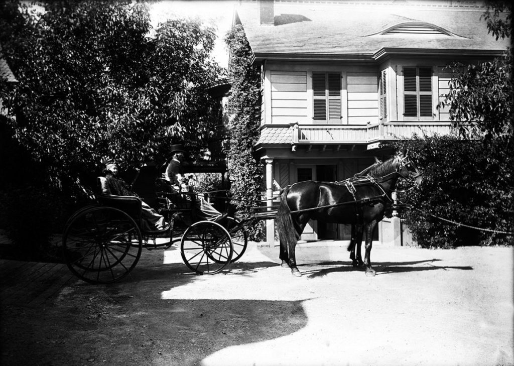 A horse and carriage bearing Sarah Winchester, outside of a house.