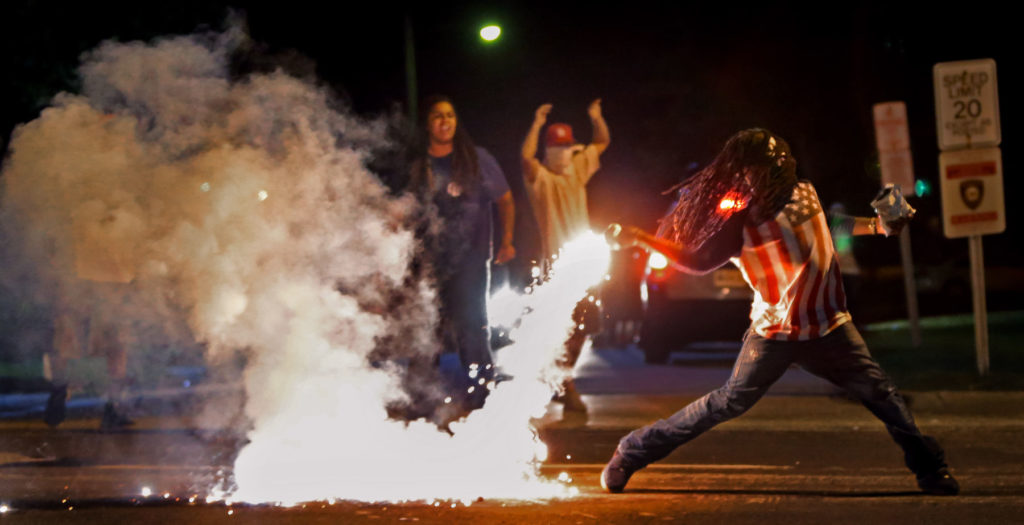 Edward Crawford, wearing an American flag T-shirt, picks up a smoking tear gas canister, and begins to throw it back toward police.