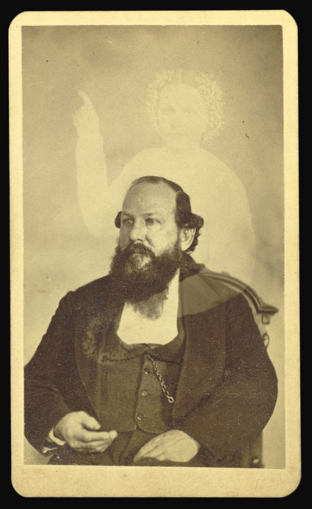 Sepia portrait photograph from the 1800s showing a man sitting in a chair with a transparent woman standing behind him, one arm wrapped around the man, the other pointed to the sky.
