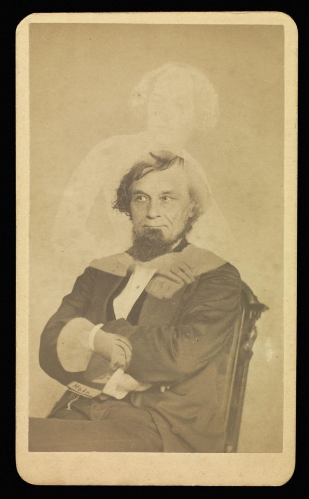 Sepia portrait photograph from the 1800s showing a man sitting in a chair, arms crossed, with a transparent woman standing behind him, arms appearing to be wrapped around the man.