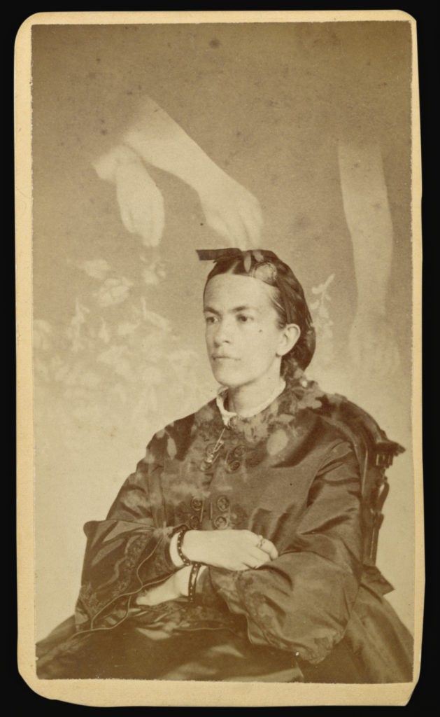 Sepia portrait photograph from the 1800s showing a woman sitting in a chair, angled and gazing beyond of the camera, with her arms crossed in her lap. Above her, three transparent arms place transparent flowers around the woman.