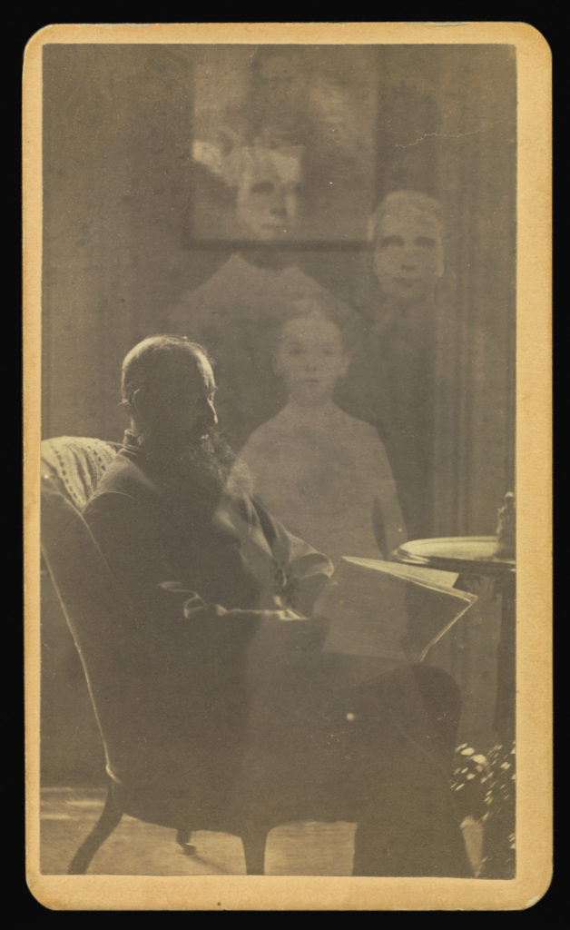 Sepia portrait photograph from the 1800s showing a man sitting in a chair reading a newspaper; next to him stand three transparent people.